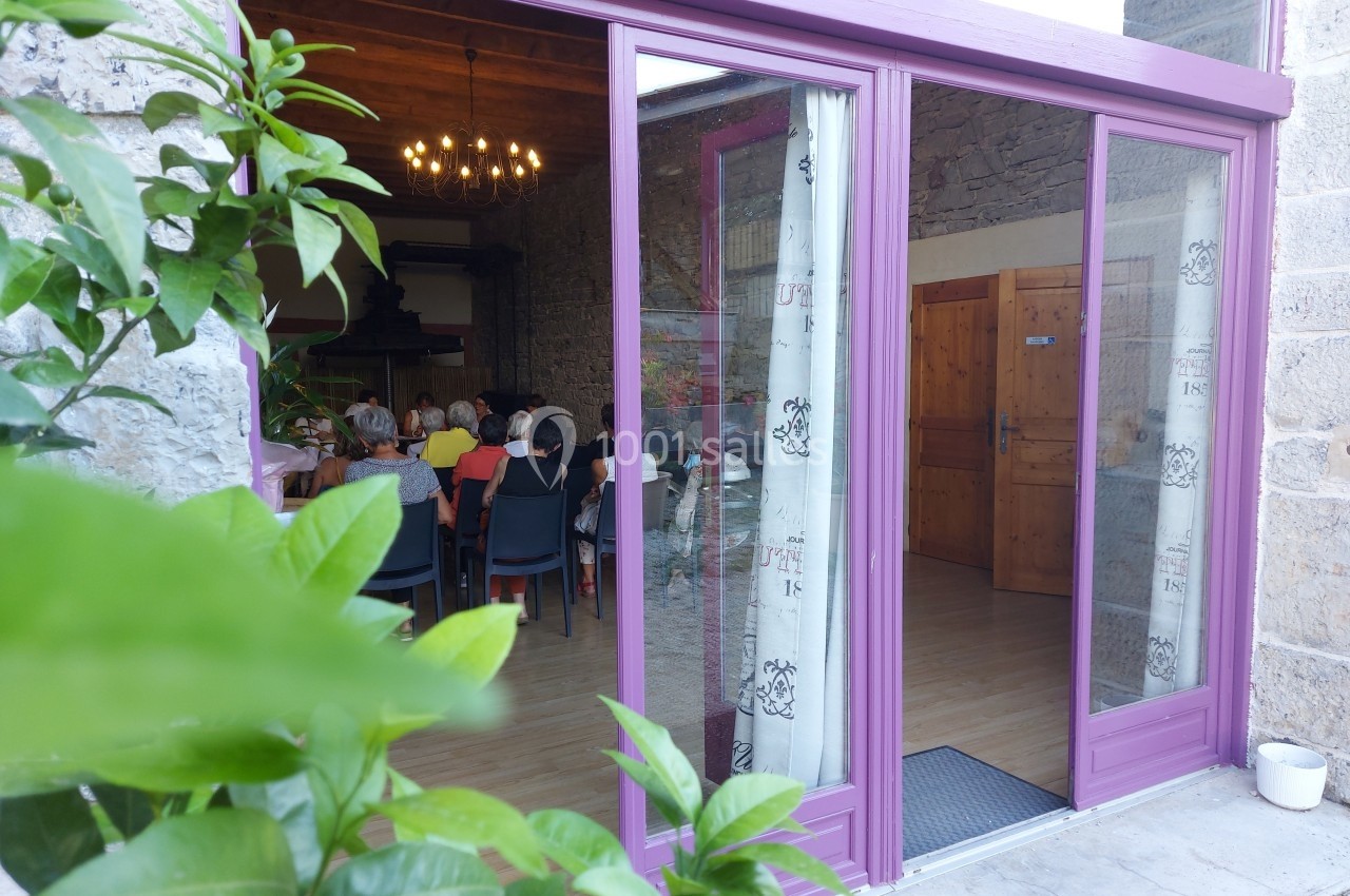 Location salle Quissac (Gard) - Le Mas de Roux #6 Vue d'une salle de réunion à travers une baie vitrée ouverte, avec des personnes assises autour de tables.