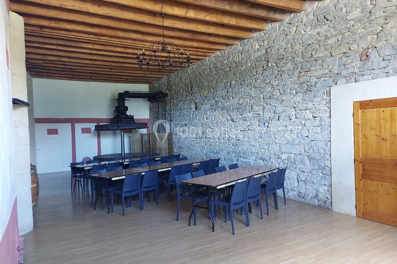 Location salle Quissac (Gard) - Le Mas de Roux #10 Salle avec des tables et chaises alignées, murs en pierre, plafond en bois avec poutres apparentes et un ancien pressoir.