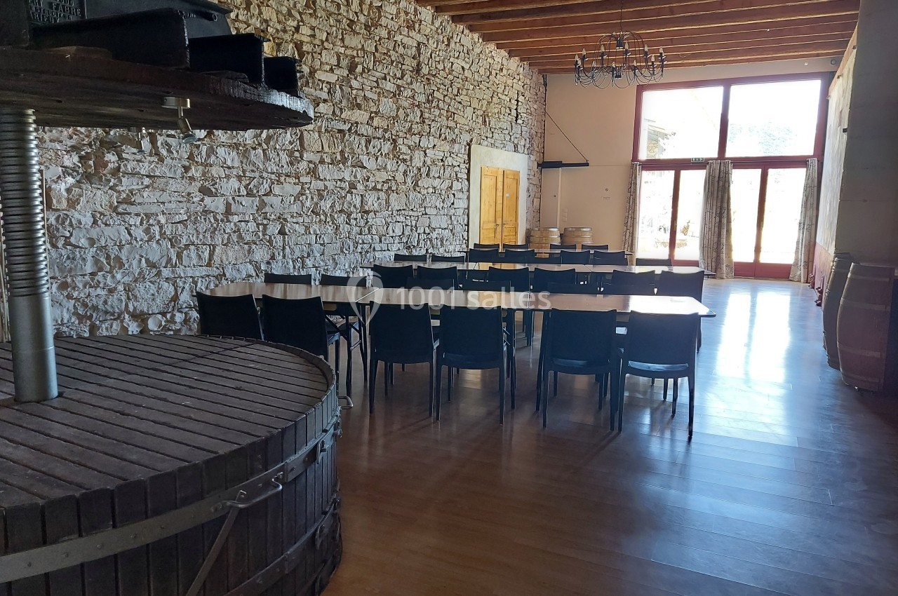 Location salle Quissac (Gard) - Le Mas de Roux #3 Salle avec murs en pierre, tables alignées avec chaises noires, grande baie vitrée et ancien pressoir en bois au premier…