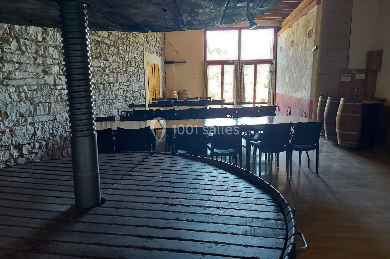 Location salle Quissac (Gard) - Le Mas de Roux #5 Salle lumineuse avec murs en pierre, tables et chaises noires, tonneaux en bois et grande presse au premier plan.