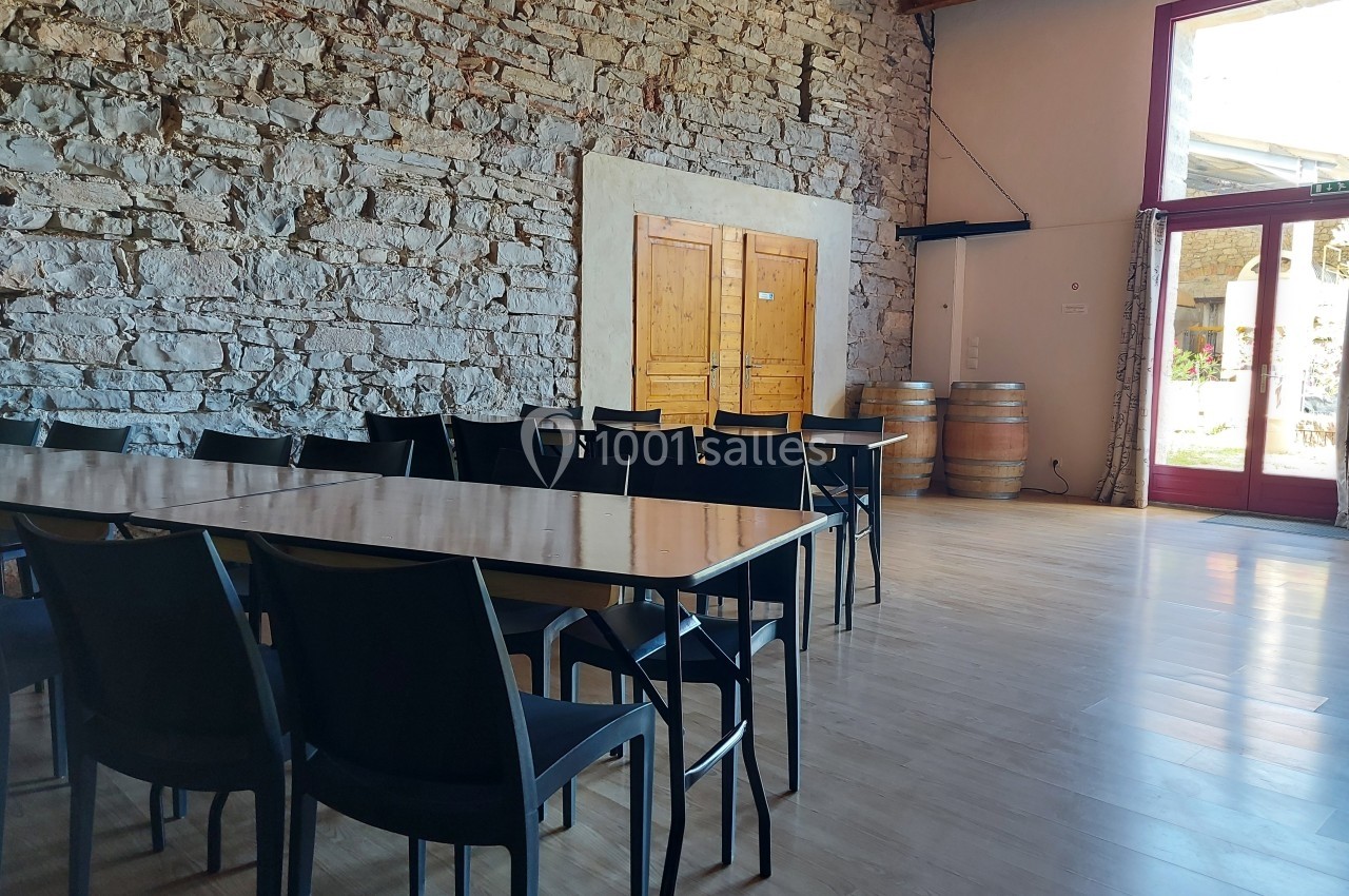 Location salle Quissac (Gard) - Le Mas de Roux #11 Salle lumineuse avec murs en pierre, tables et chaises noires, barriques en bois et grande porte vitrée.