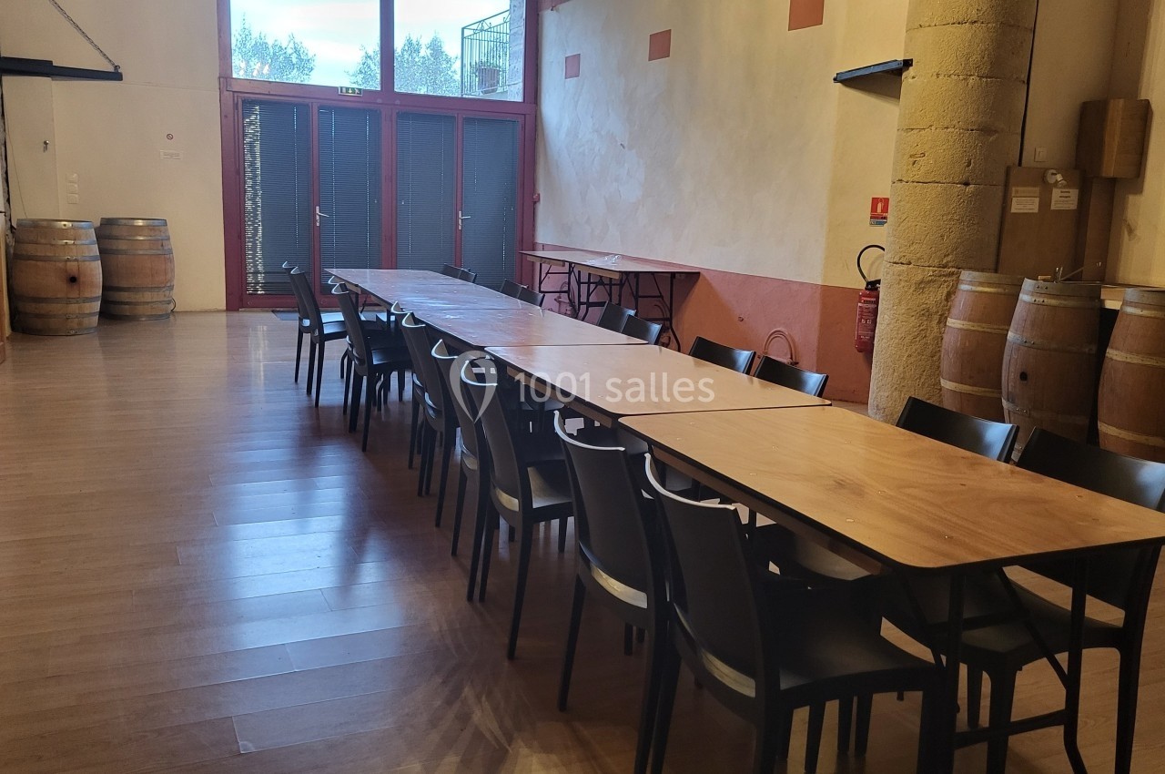 Location salle Quissac (Gard) - Le Mas de Roux #12 Salle avec une longue table en bois entourée de chaises noires, des tonneaux et un mur en pierre en arrière-plan.