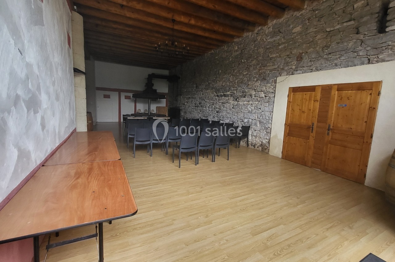 Location salle Quissac (Gard) - Le Mas de Roux #4 Salle avec murs en pierre et plafond en bois, équipée de chaises noires, tables en bois et tonneaux dans un coin.
