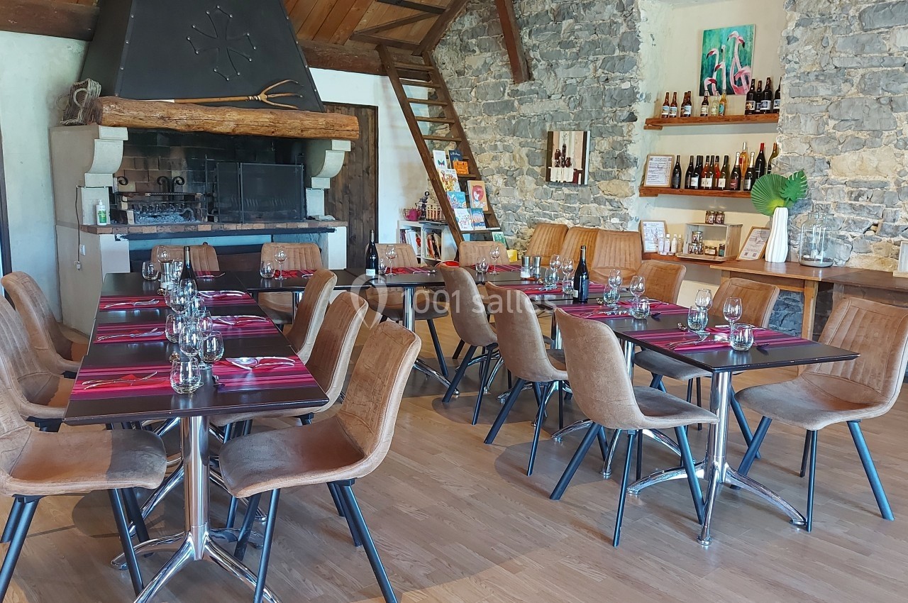 Location salle Quissac (Gard) - Le Mas de Roux #15 Salle de restaurant avec tables dressées, chaises en cuir, cheminée en pierre et étagères garnies de bouteilles.