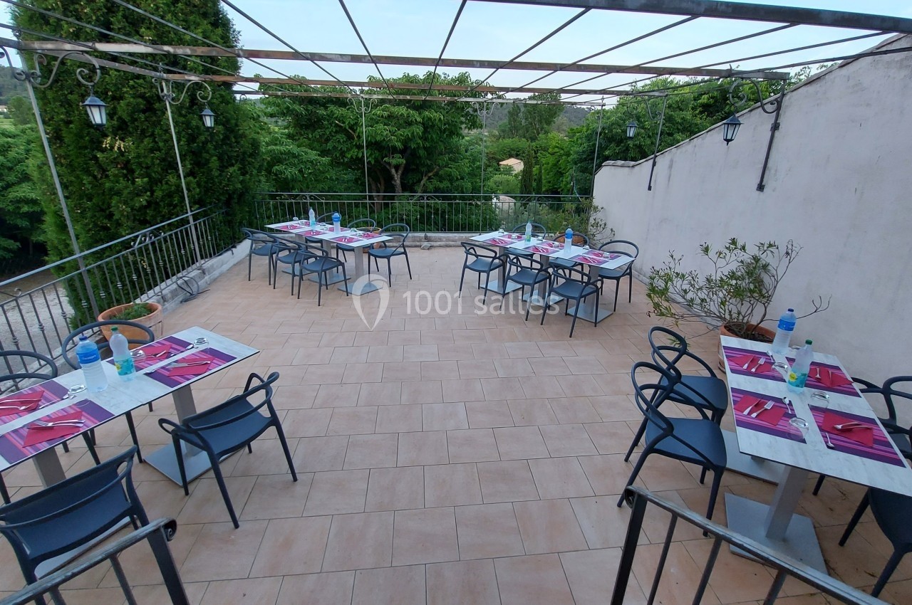 Location salle Quissac (Gard) - Le Mas de Roux #7 Terrasse extérieure avec tables dressées, chaises noires et vue sur des arbres et collines verdoyantes.