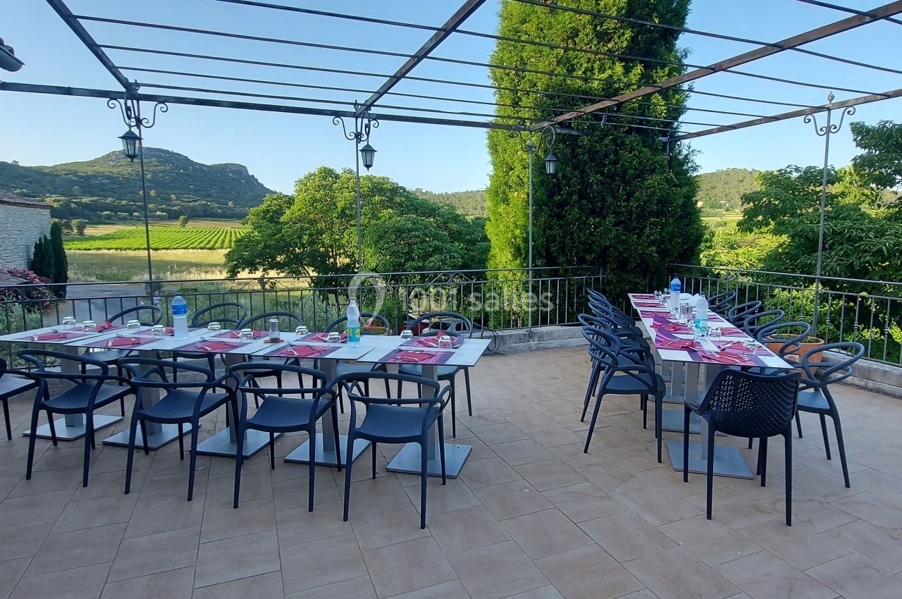 Location salle Quissac (Gard) - Le Mas de Roux #8 Terrasse aménagée avec deux grandes tables dressées, entourée de verdure et offrant une vue sur des collines.