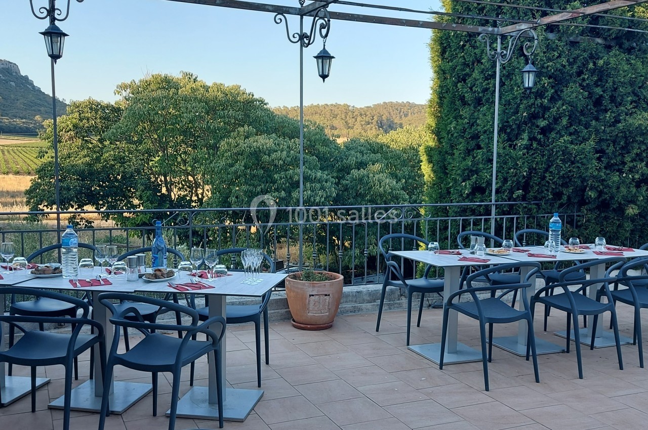 Location salle Quissac (Gard) - Le Mas de Roux #9 Terrasse extérieure avec tables dressées, chaises noires et vue sur un paysage de campagne avec collines et végétation.