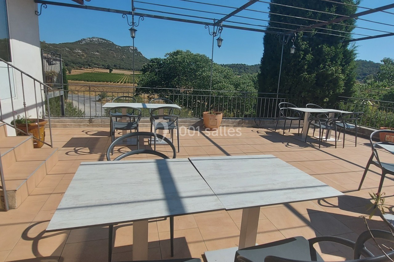 Location salle Quissac (Gard) - Le Mas de Roux #16 Terrasse ensoleillée avec tables et chaises, entourée de verdure et offrant une vue sur des collines et des vignes.