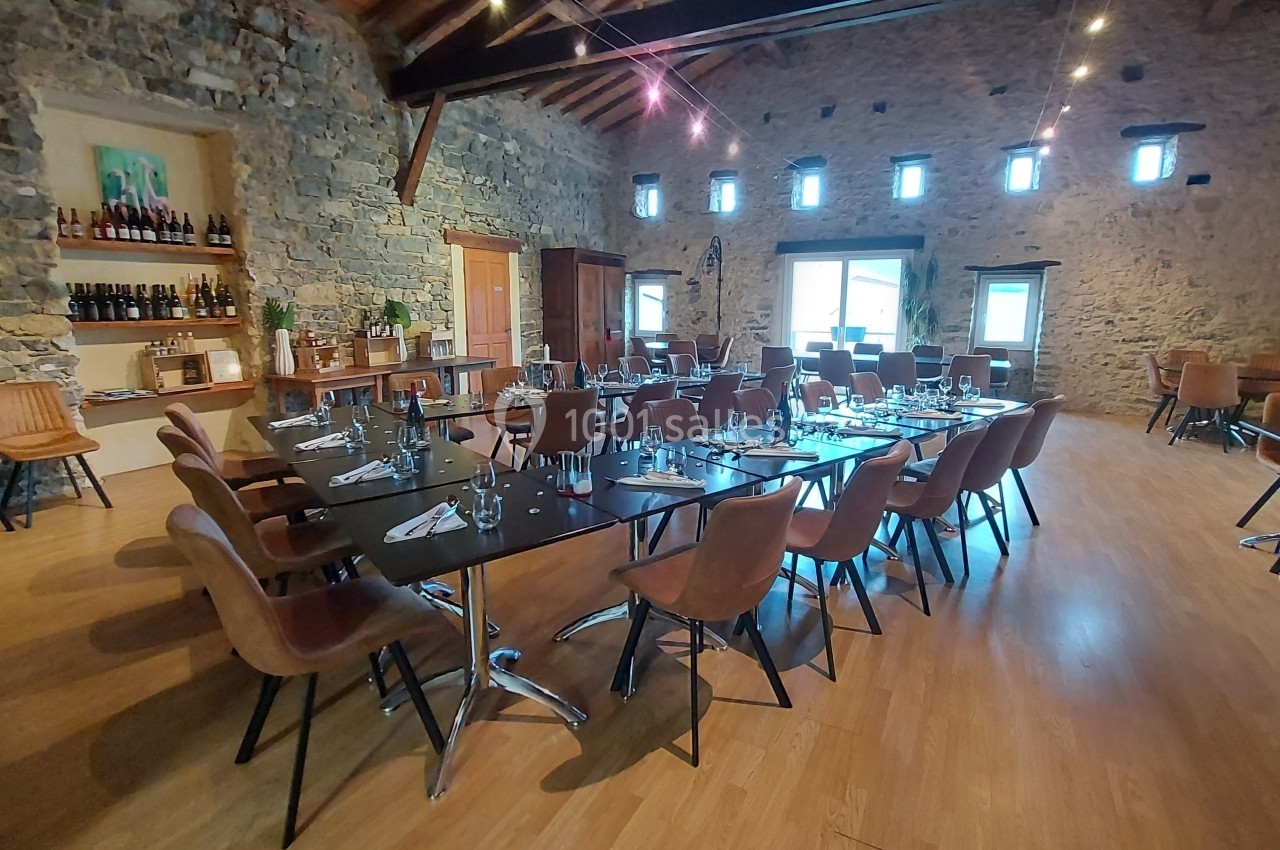 Location salle Quissac (Gard) - Le Mas de Roux #18 Salle en pierre avec tables dressées, chaises en cuir, étagères de bouteilles et lumière naturelle provenant des fenêtres.