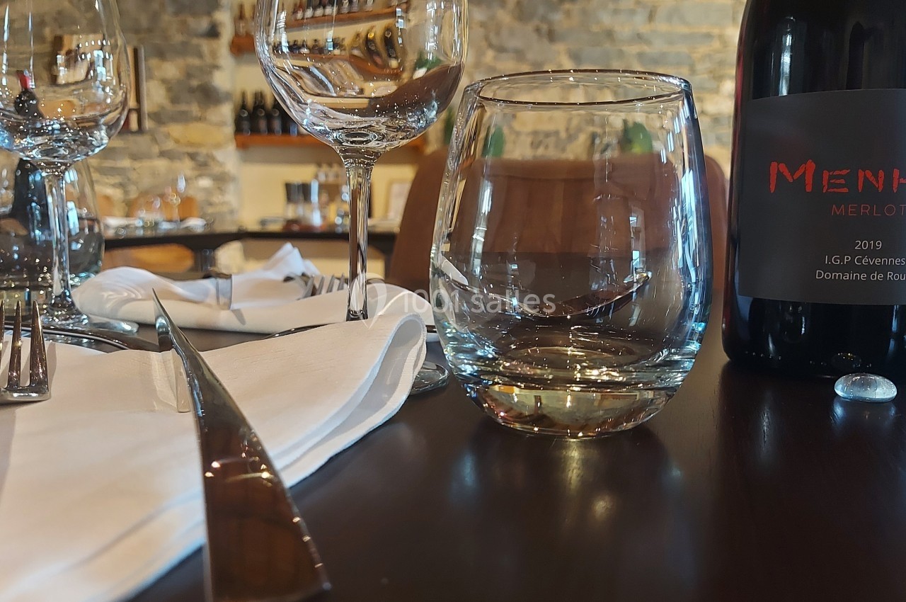 Location salle Quissac (Gard) - Le Mas de Roux #19 Bouteille de vin rouge, verres et couverts disposés sur une table en bois dans un cadre de restaurant en pierre.