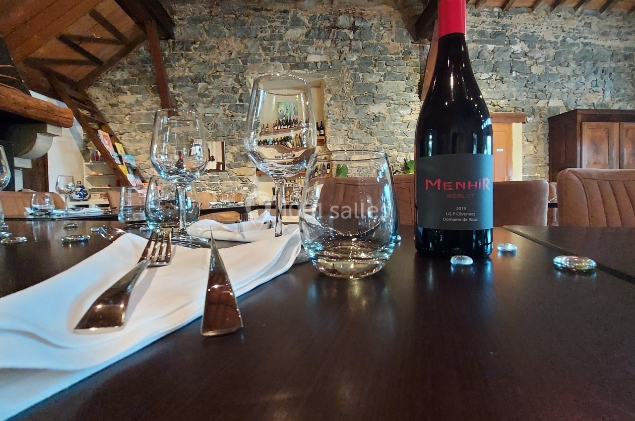 Location salle Quissac (Gard) - Le Mas de Roux #20 Table dressée avec des verres, couverts et une bouteille de vin rouge dans une salle à murs en pierre apparente.