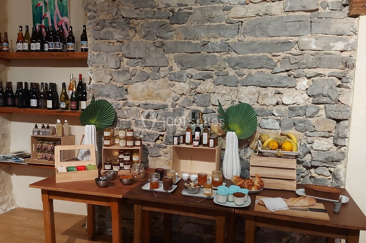 Location salle Quissac (Gard) - Le Mas de Roux #21 Table avec produits locaux : confitures, miel, pain, viennoiseries, fruits, boissons et décorations végétales devant un mur…