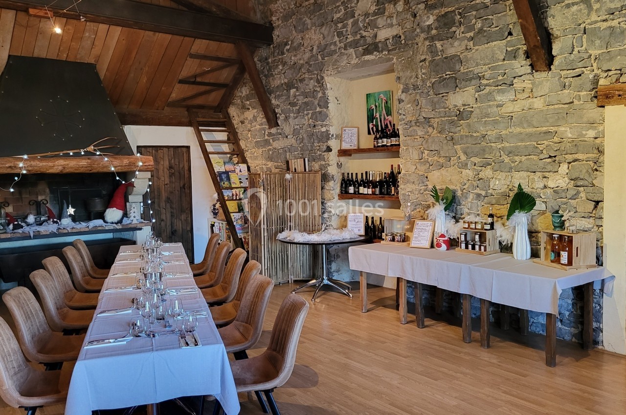 Location salle Quissac (Gard) - Le Mas de Roux #25 Salle rustique avec murs en pierre, grande table dressée, cheminée décorée et tables présentant des produits locaux.
