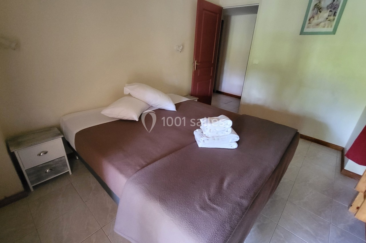 Location salle Quissac (Gard) - Le Mas de Roux #27 Lit double avec draps bruns, oreillers blancs et serviettes pliées, dans une chambre lumineuse avec porte rouge ouverte.