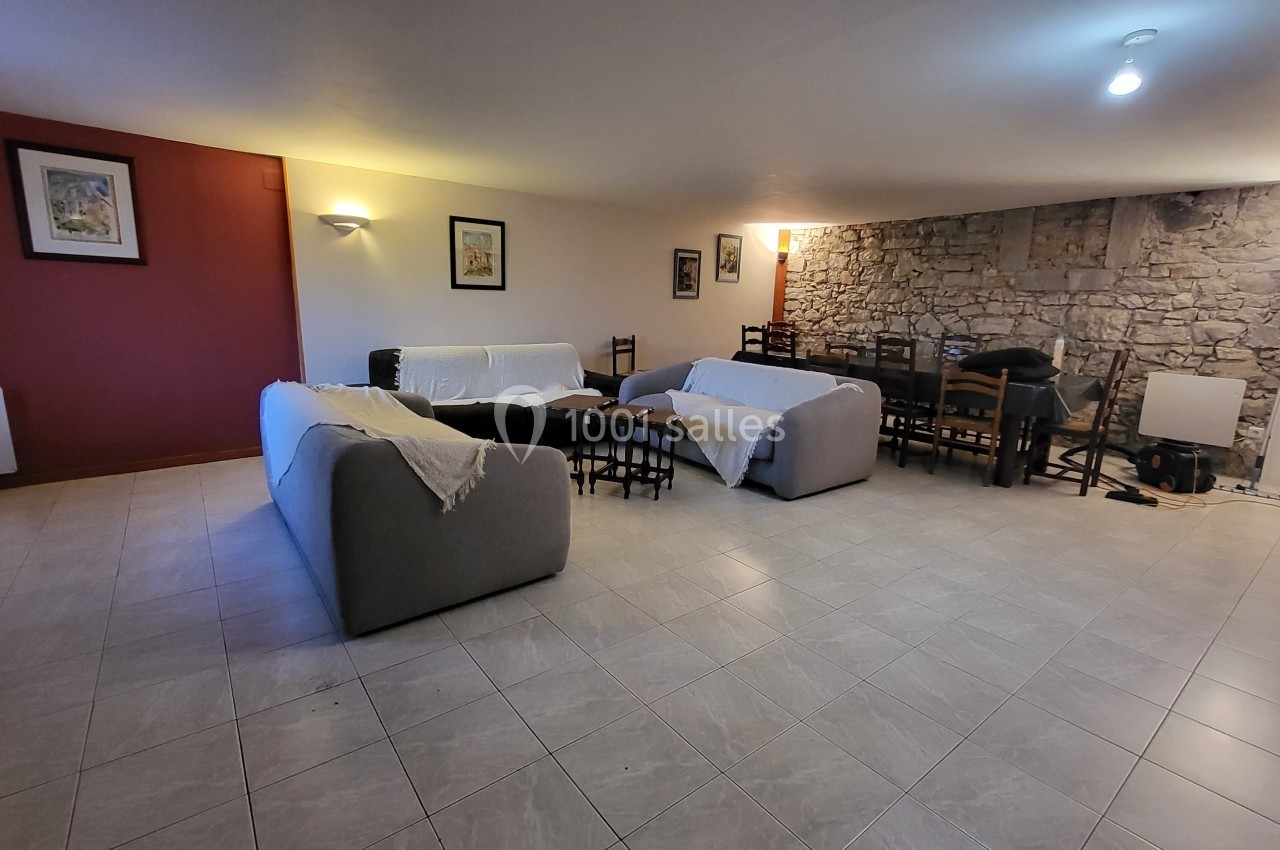 Location salle Quissac (Gard) - Le Mas de Roux #29 Salle spacieuse avec canapés recouverts de plaids, tables et chaises en bois, mur en pierre et éclairage doux.