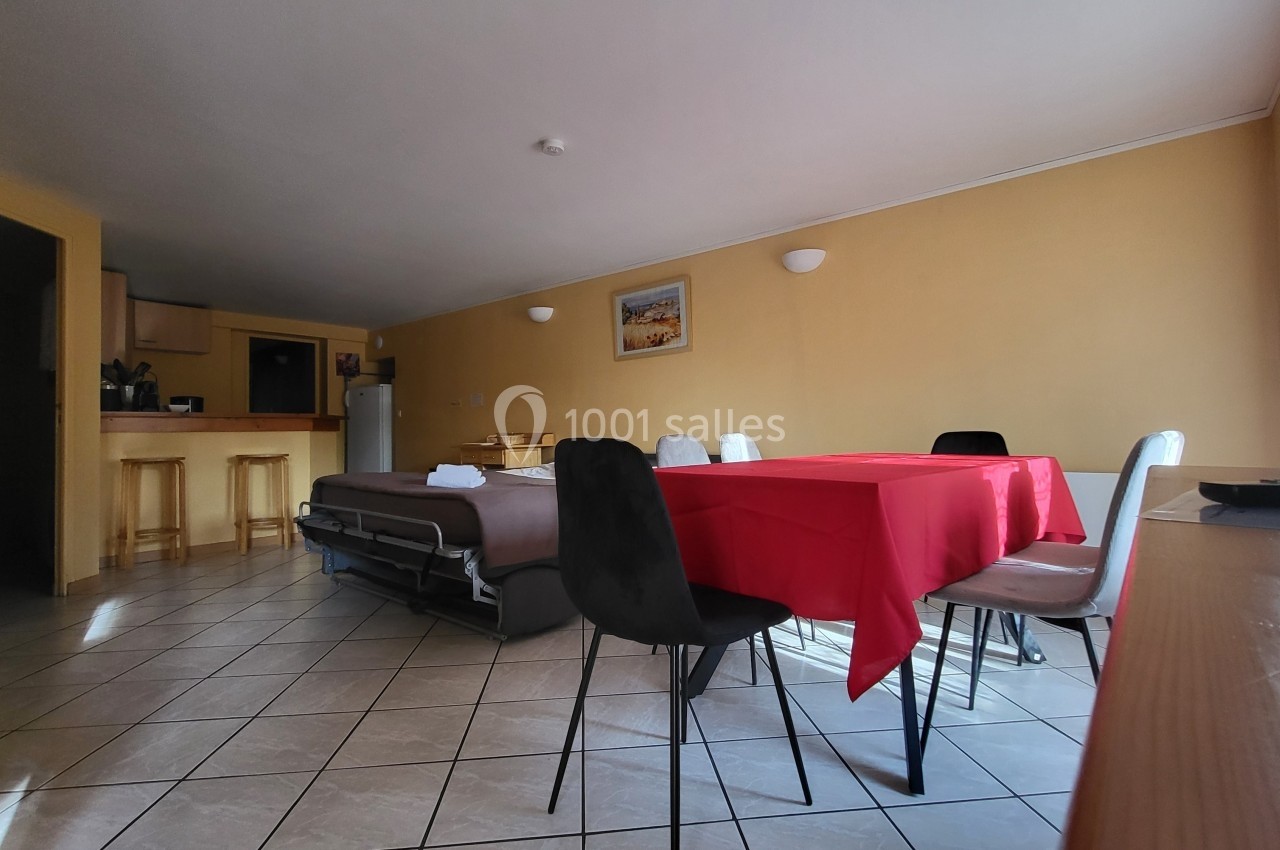 Location salle Quissac (Gard) - Le Mas de Roux #31 Salle à manger avec table recouverte d'une nappe rouge, chaises noires et blanches, cuisine ouverte et canapé-lit en arrière…