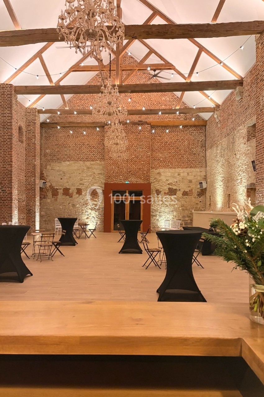 Salle en briques avec poutres apparentes, tables hautes noires et éclairage chaleureux, vue depuis un comptoir en bois.