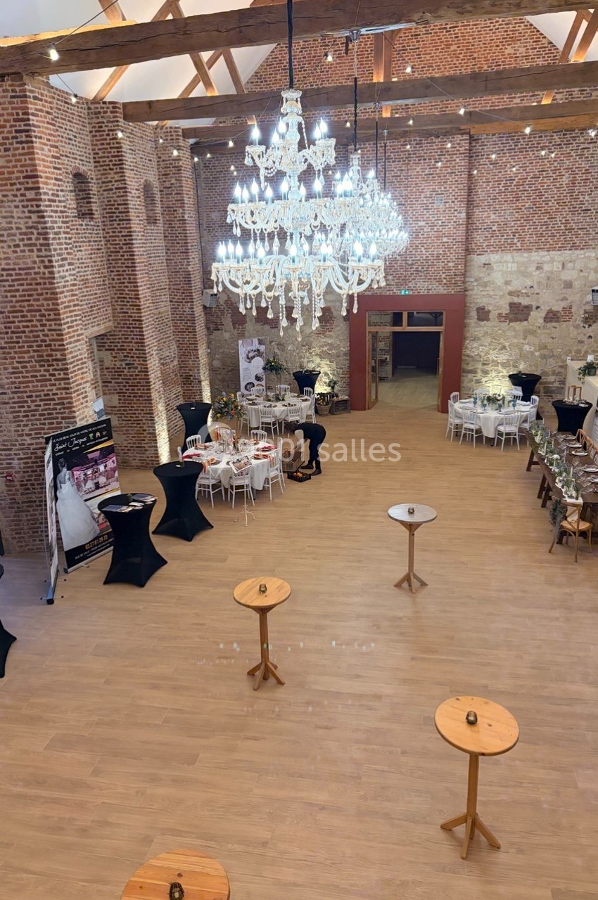 Salle de réception spacieuse avec murs en briques, parquet clair, tables hautes et un grand lustre central.