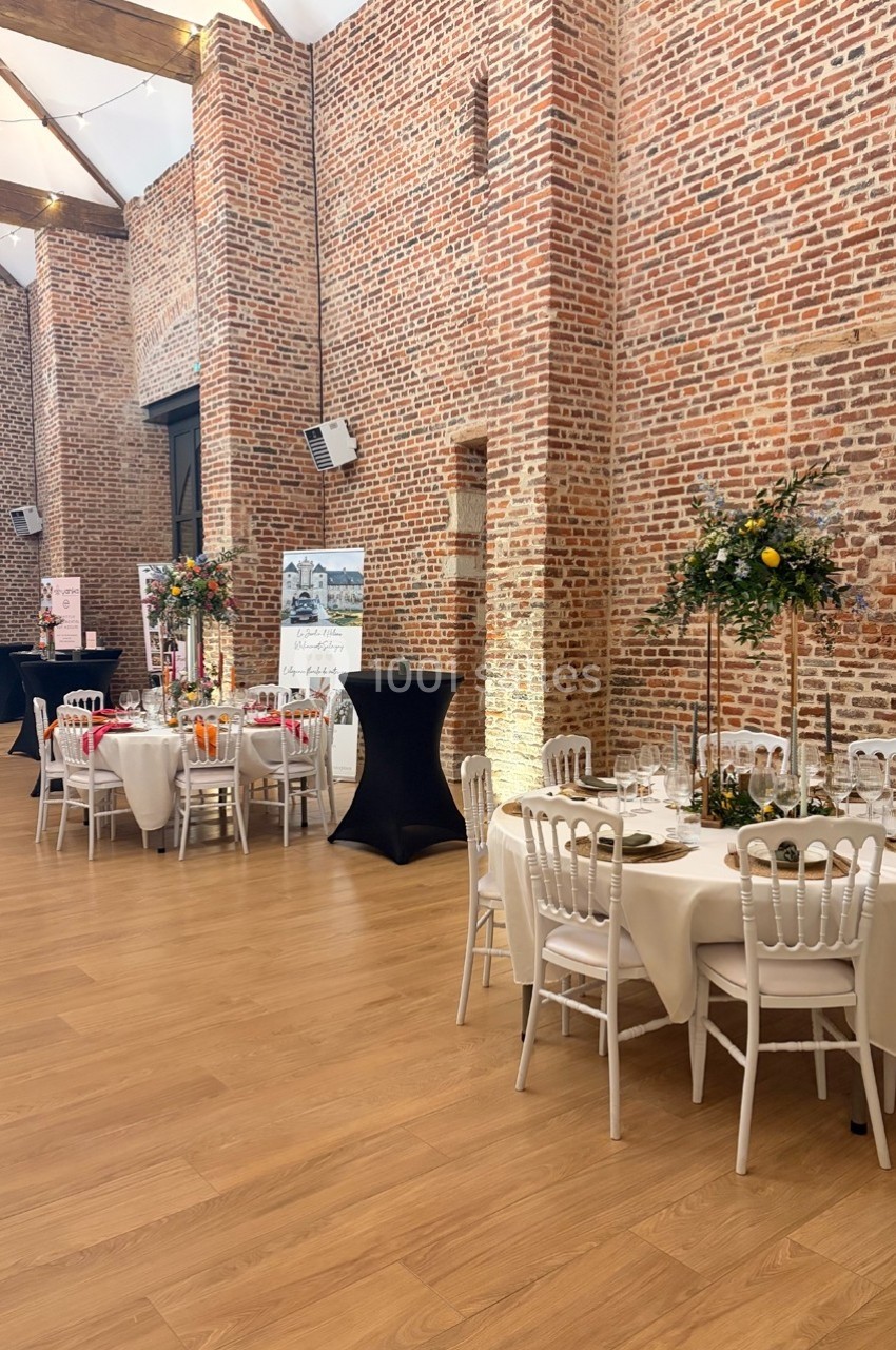Salle de réception avec murs en briques, tables rondes décorées et chaises blanches sur un sol en bois clair.