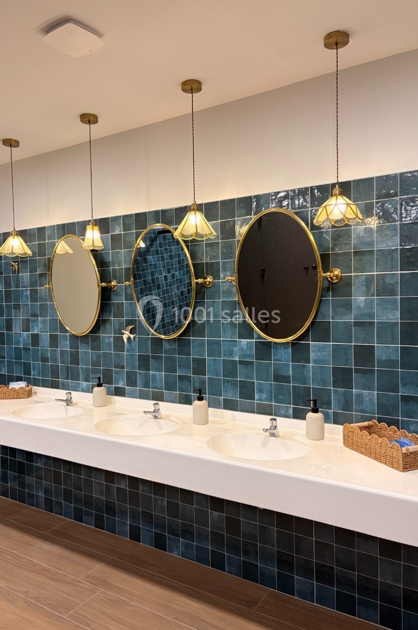 Miroirs ronds dorés et lavabos blancs alignés sur un mur carrelé bleu, éclairés par des suspensions décoratives.
