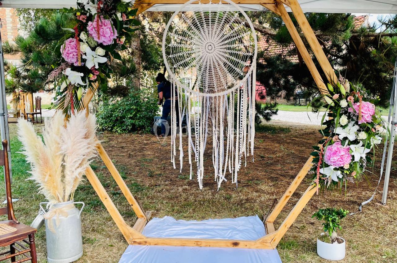 Structure hexagonale en bois décorée de fleurs, d'un attrape-rêves et de pampas, installée en extérieur.