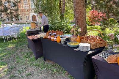 Miniature Brunch dans la cour intérieure du Château  Pièce rustique avec murs en pierre, plafond en bois, réservoir métallique rouge et mobilier simple.