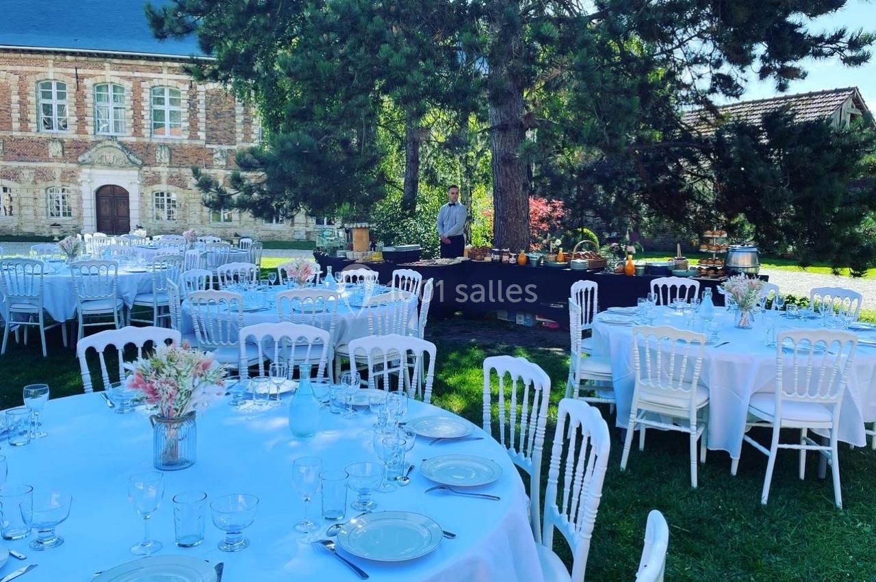 Tables dressées pour un événement en plein air dans un jardin, avec un buffet et un bâtiment en arrière-plan.
