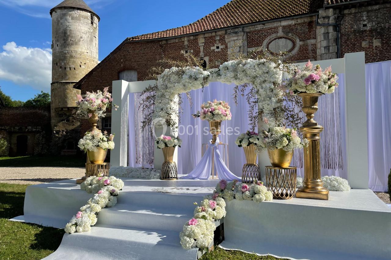Décor de mariage en extérieur avec arche florale blanche et rose, colonnes dorées et fond de bâtiment en briques.