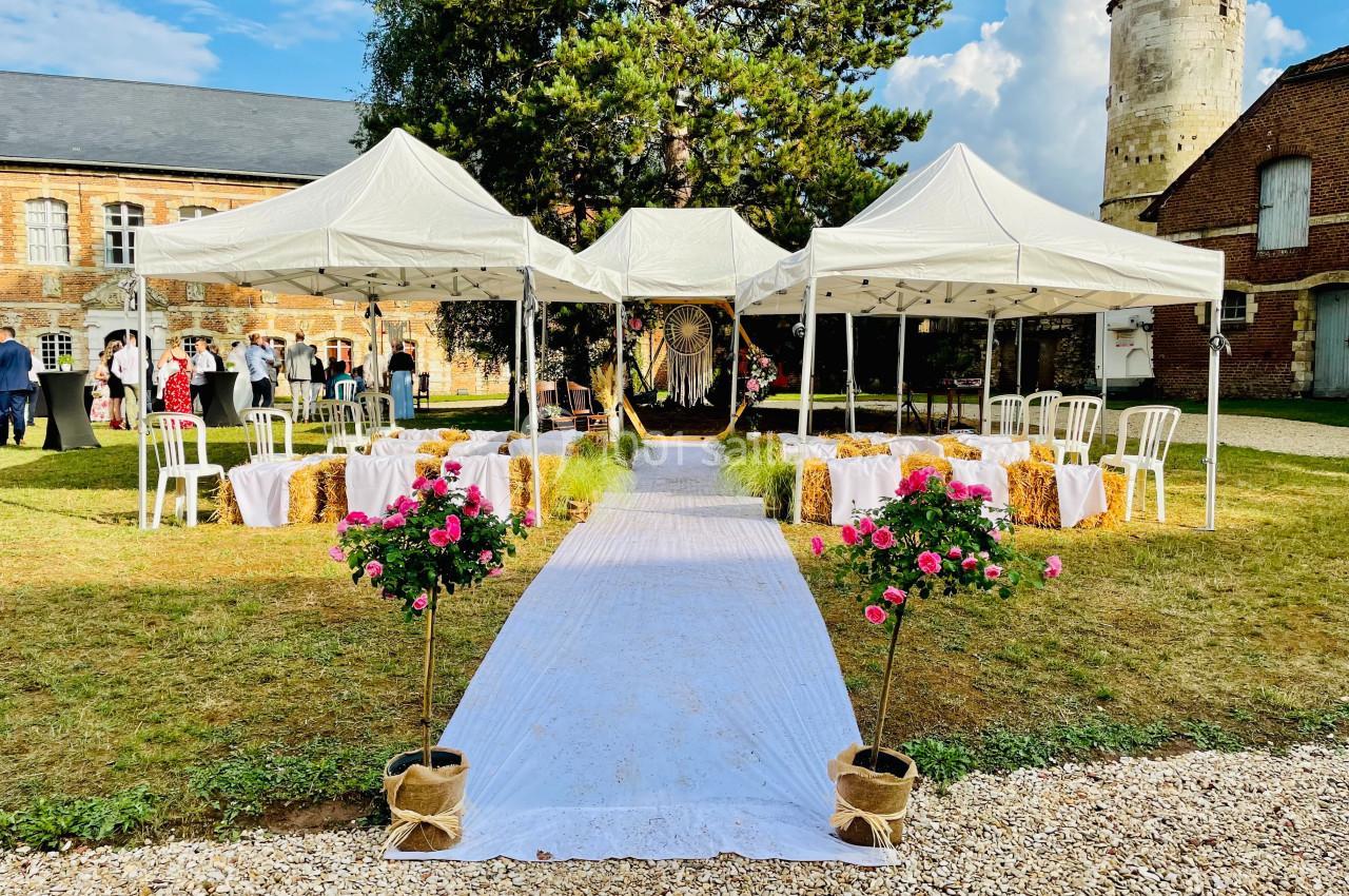 Allée centrale bordée de fleurs menant à une cérémonie en plein air sous des tentes blanches dans un cadre champêtre.