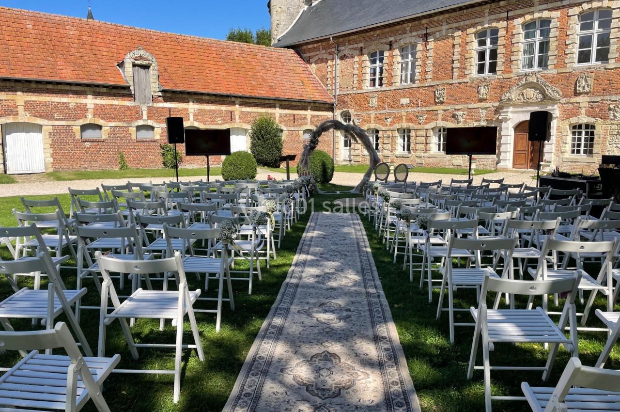 Chaises blanches disposées en rangées face à une arche décorative dans une cour extérieure pavée d'herbe.