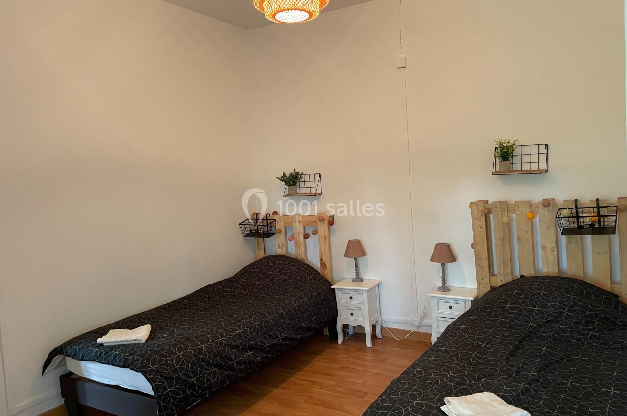 Deux lits simples avec linge de lit sombre, tables de chevet, lampes et décoration murale dans une chambre lumineuse.