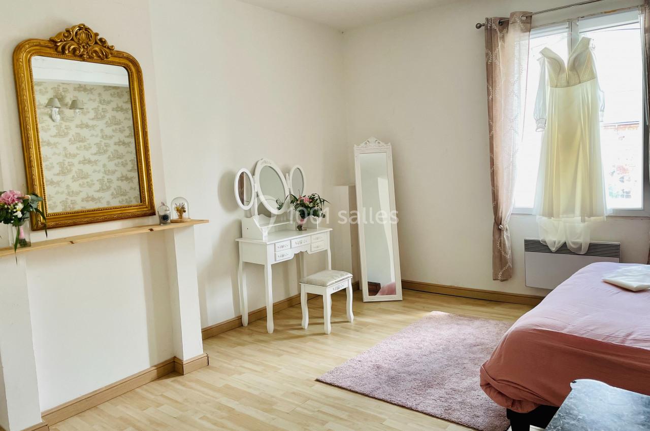 Chambre lumineuse avec coiffeuse blanche, miroir doré, robe suspendue à la fenêtre et lit avec couverture rose.