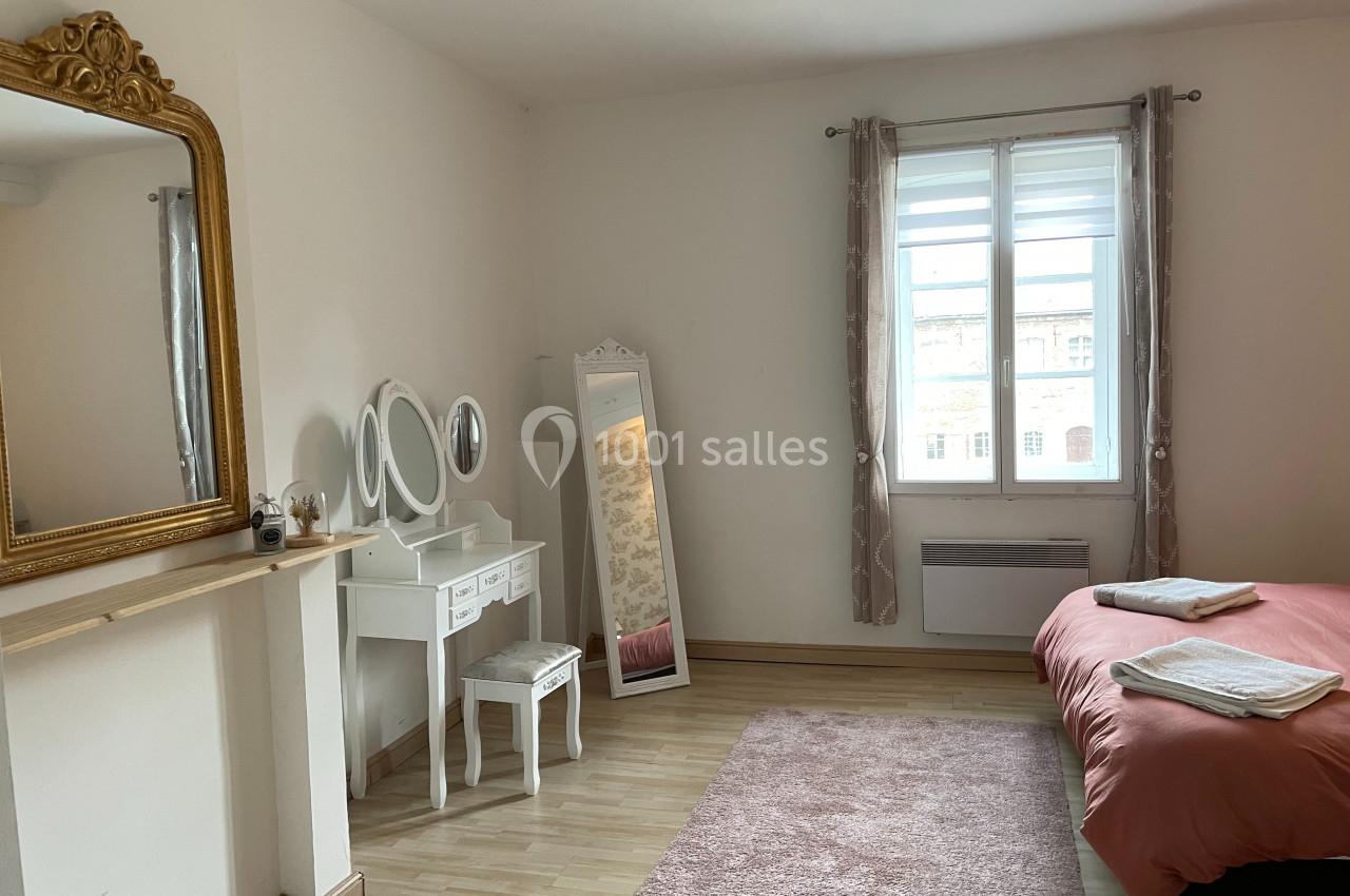 Chambre lumineuse avec lit double, coiffeuse blanche, miroir doré et grande fenêtre avec rideaux roses.