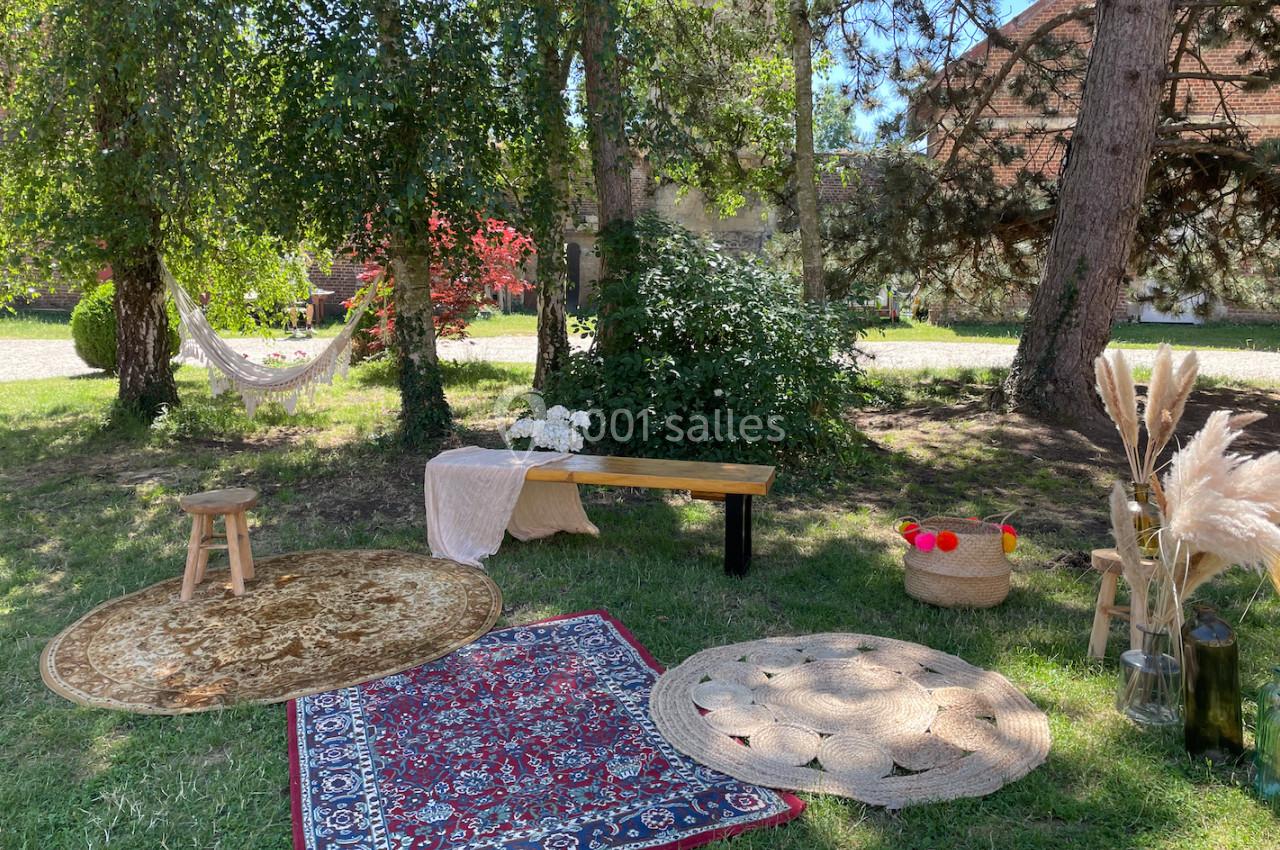 Espace extérieur aménagé avec tapis, banc, tabouret, hamac et décorations végétales sous des arbres.
