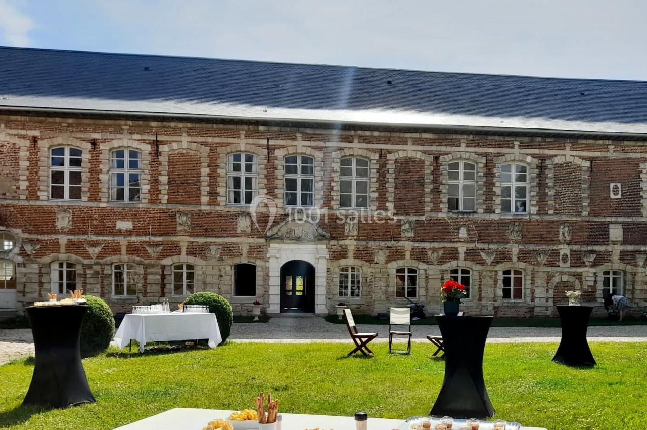 Façade en briques d'un bâtiment ancien avec tables dressées et chaises sur une pelouse ensoleillée.