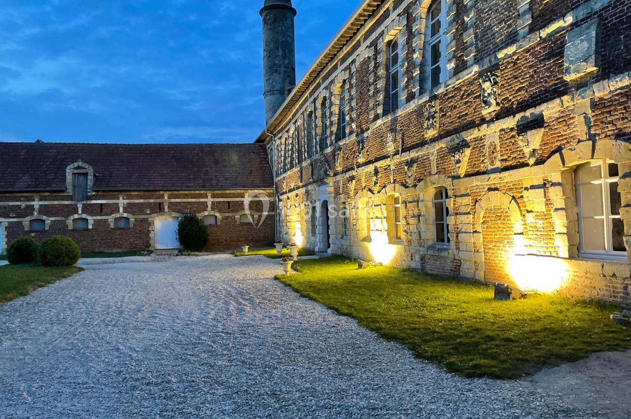 Cour pavée d'un bâtiment en briques anciennes, éclairé par des lumières au crépuscule, avec une cheminée en arrière-plan.