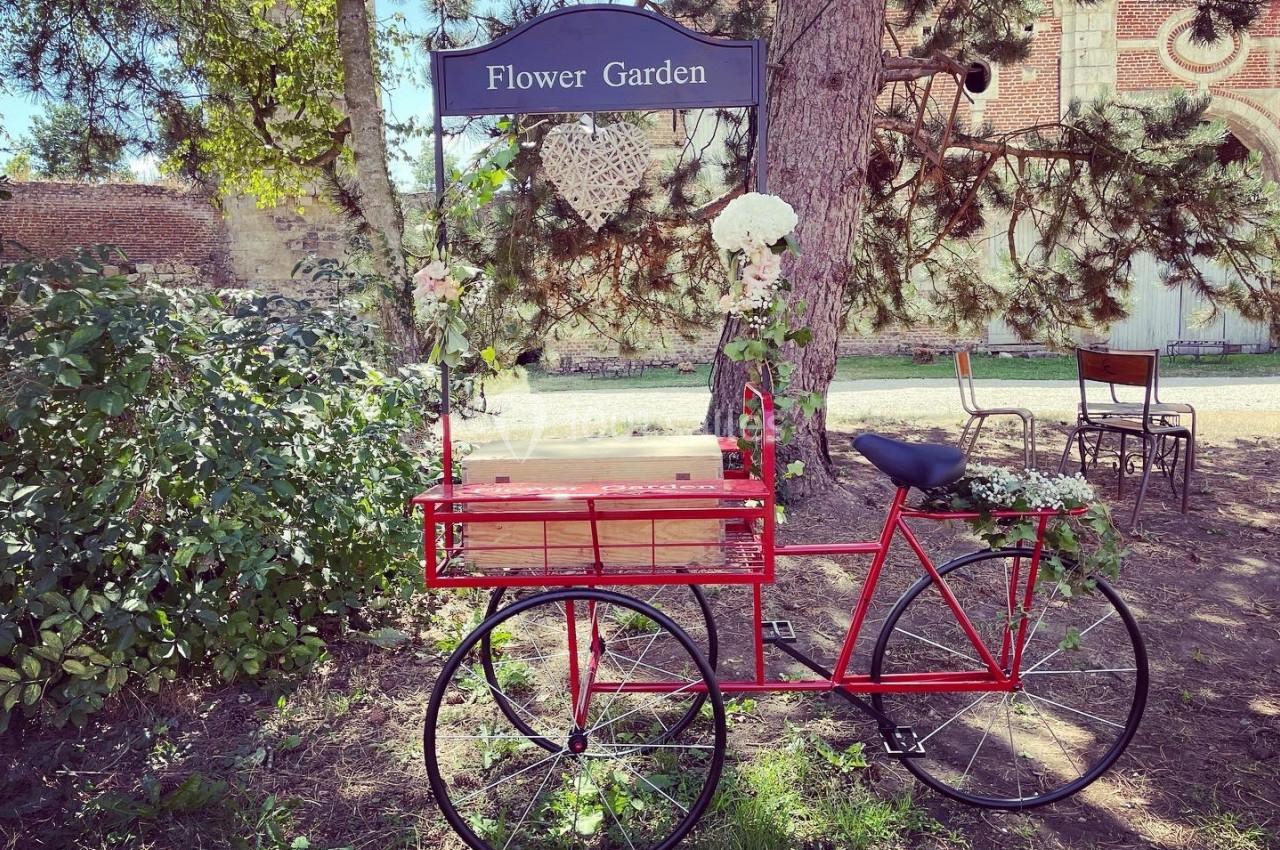 Vélo rouge décoratif avec des fleurs blanches, placé sous un panneau ’Flower Garden’ dans un cadre extérieur verdoyant.