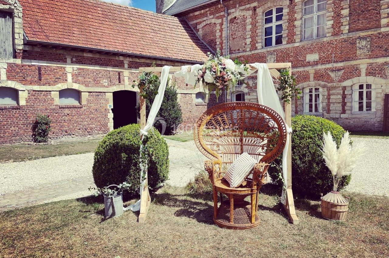 Fauteuil en rotin décoré sous une arche fleurie, installé dans une cour pavée devant un bâtiment en briques.