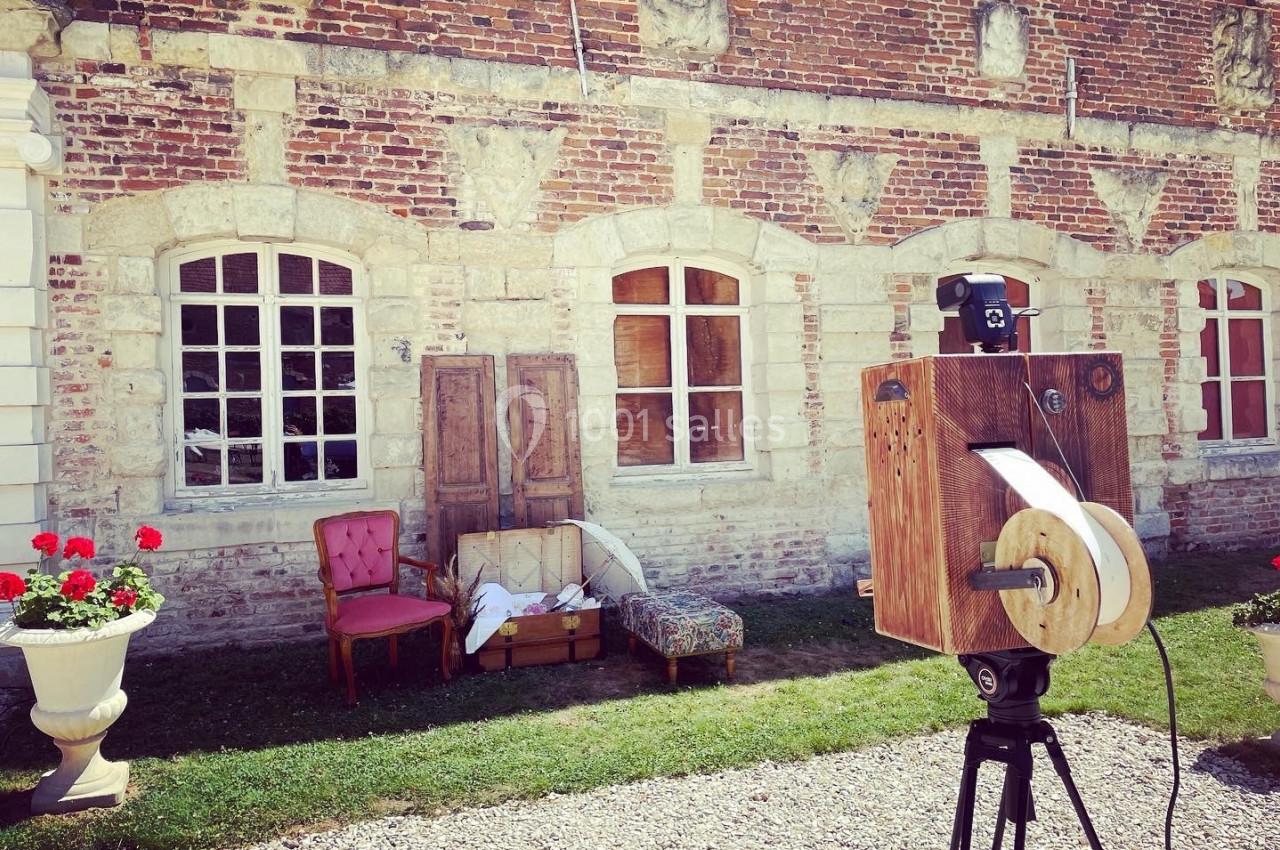 Appareil photo vintage sur trépied devant une façade en briques avec fenêtres, fauteuils et valises en arrière-plan.
