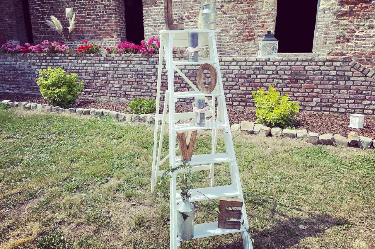 Échelle blanche décorative avec des objets et des lettres formant ’LOVE’, posée sur une pelouse devant un mur en briques.