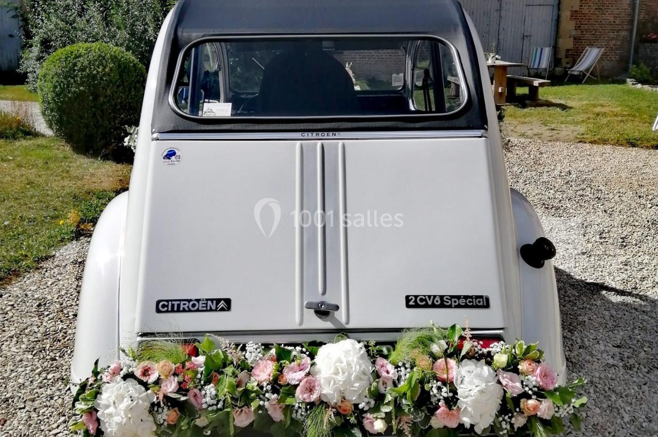 Vue arrière d'une Citroën 2CV blanche décorée de fleurs, stationnée sur un sol gravillonné devant un bâtiment en briques.