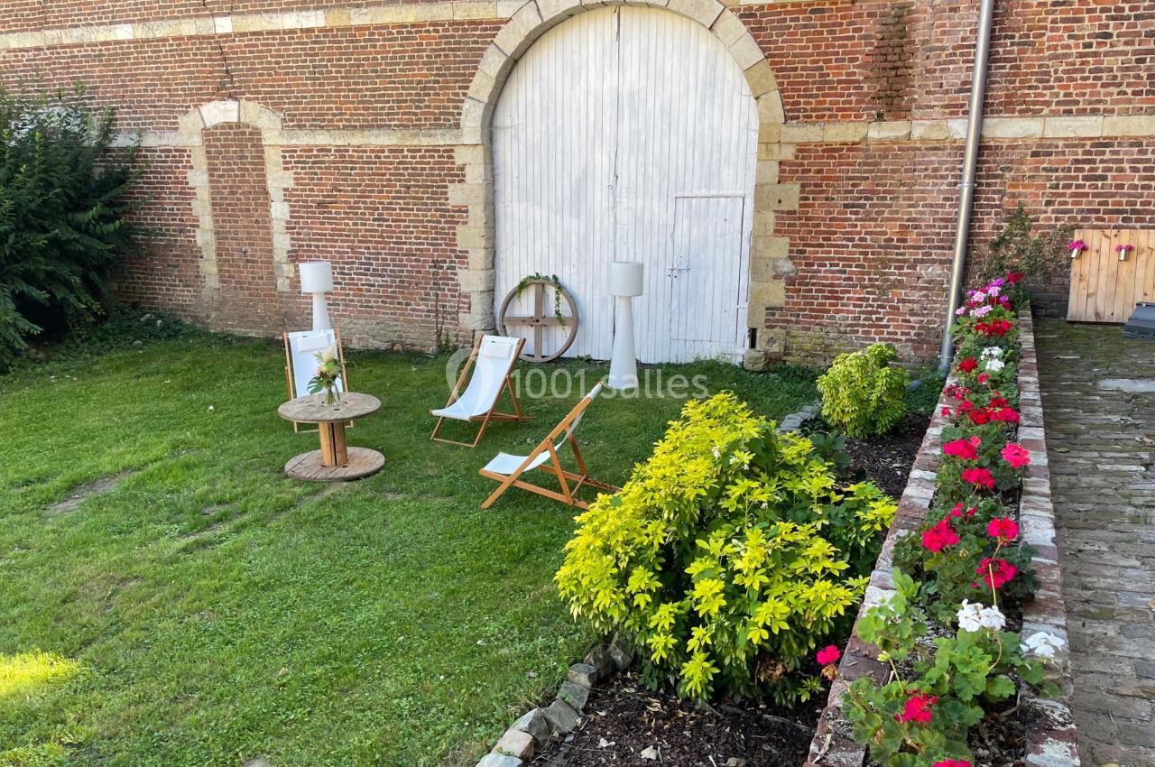 Deux chaises longues et une table basse en bois sur une pelouse près d'une porte en arc entourée de fleurs.