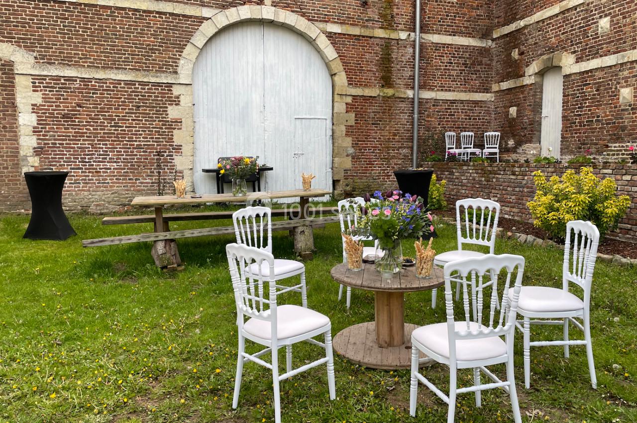 Mobilier de jardin avec chaises blanches et tables en bois disposés sur une pelouse devant un bâtiment en briques.