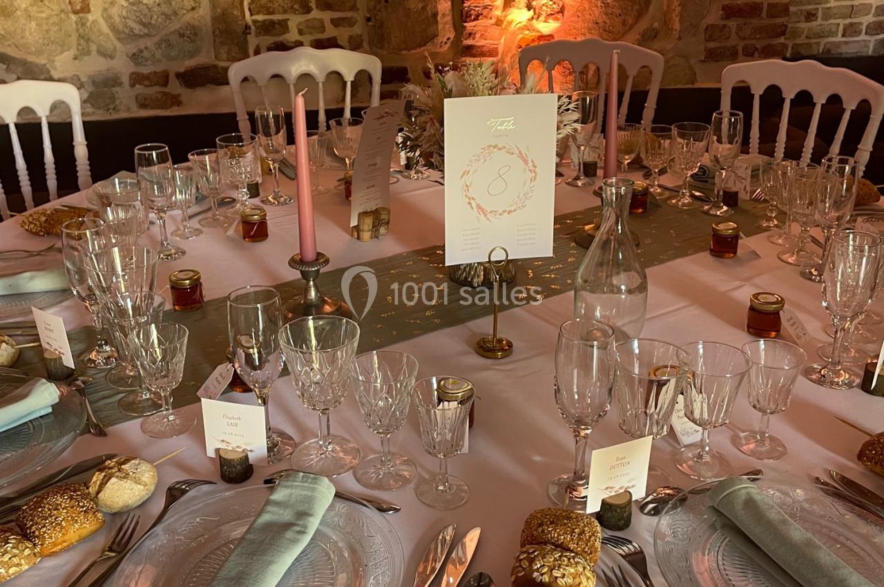 Table élégamment dressée avec nappes blanches, couverts, verres, bougies et décorations dans une salle en pierre apparente.
