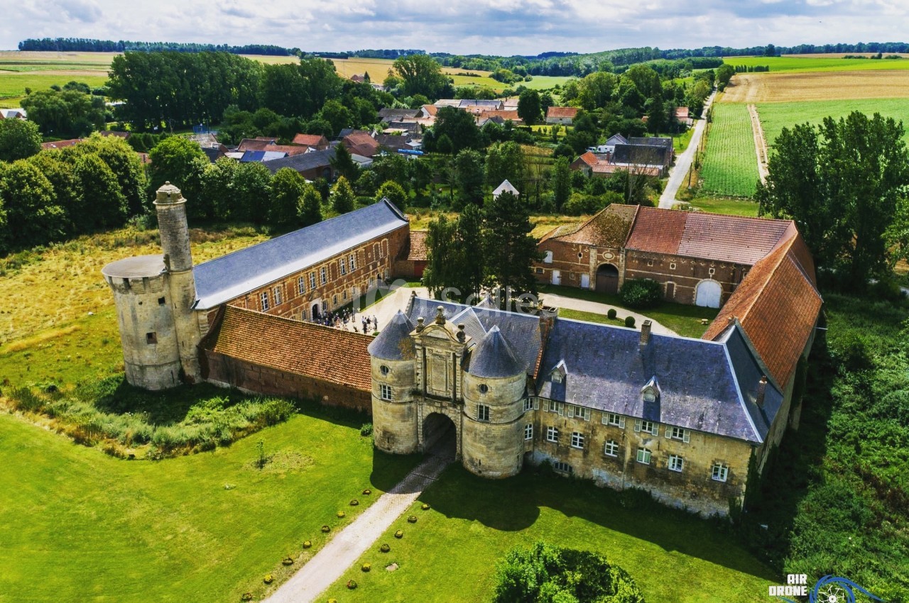 Vue aérienne d'un château historique entouré de verdure, avec des bâtiments annexes et un village en arrière-plan.