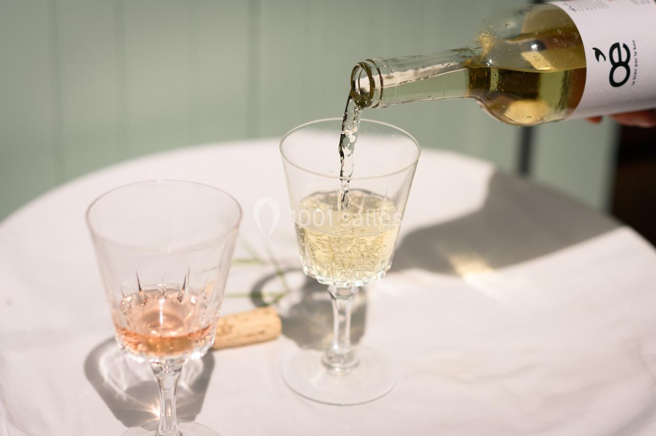 Une bouteille de vin blanc est versée dans un verre sur une table blanche, à côté d'un verre de vin rosé et d'un bouchon.