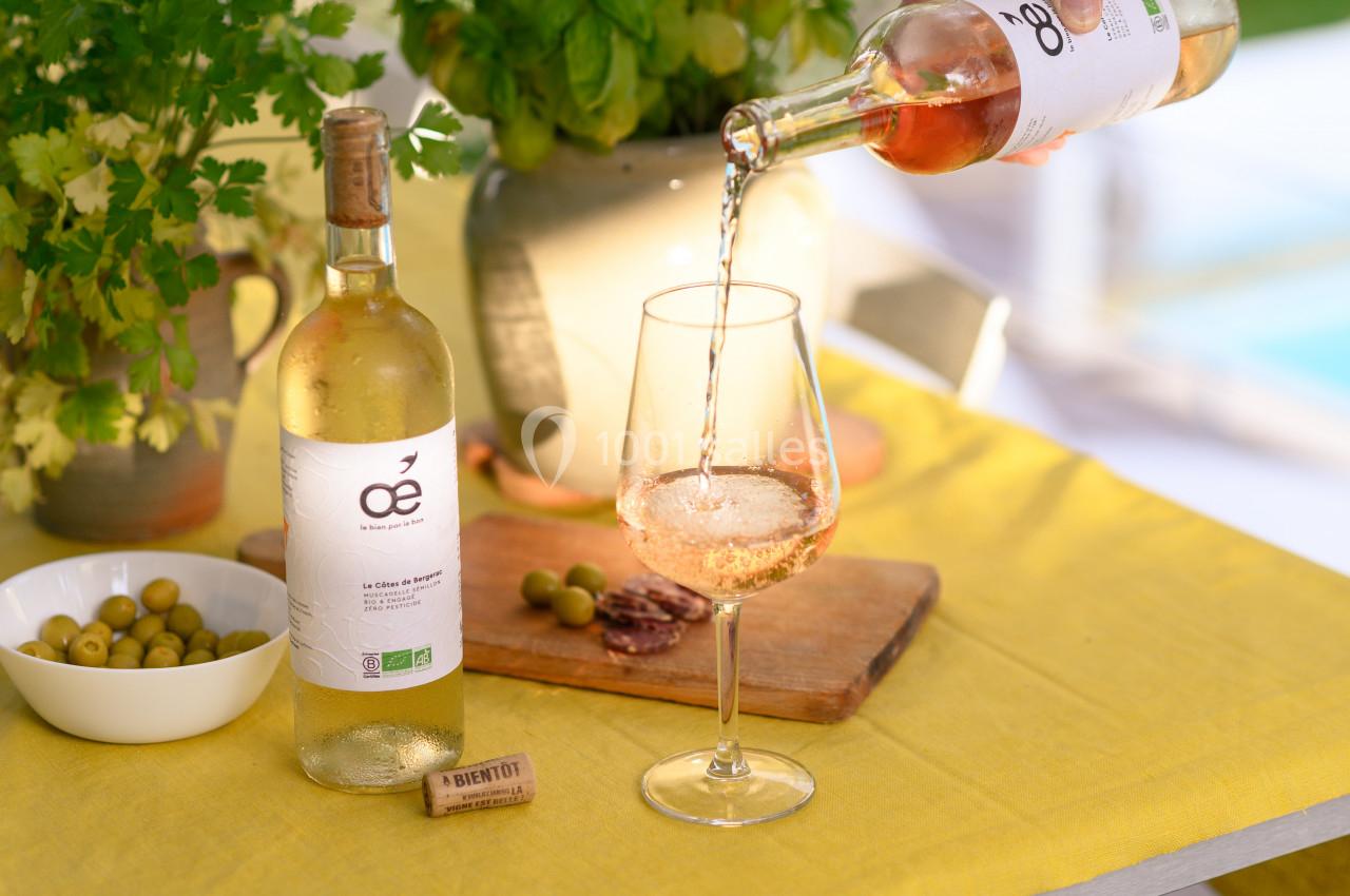 Une bouteille de vin blanc biologique est servie dans un verre sur une table avec des olives et des herbes en arrière-plan.