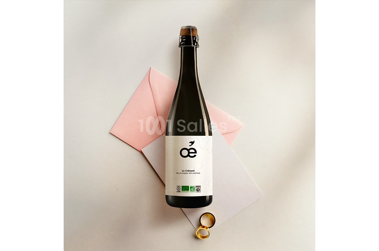 Bouteille de vin pétillant posée sur des enveloppes roses et blanches, avec deux alliances dorées à côté.