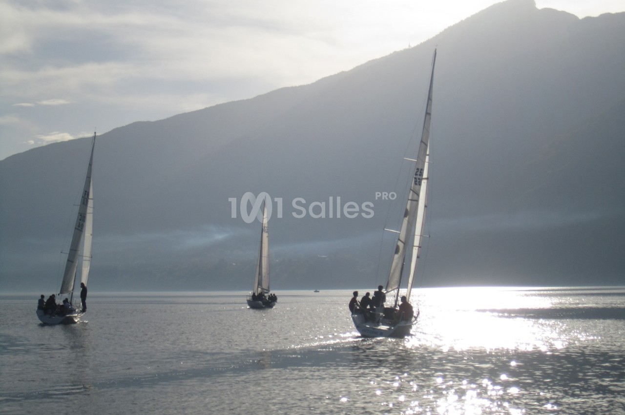 Trois voiliers naviguent sur un lac calme au lever du soleil, avec des montagnes en arrière-plan.