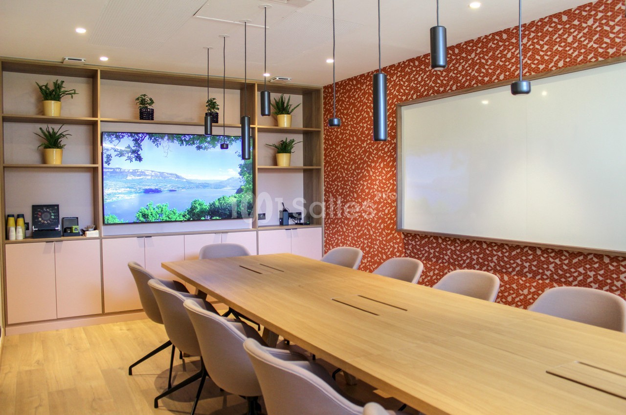Salle de réunion moderne avec grande table en bois, écran mural, tableau blanc et étagères décorées de plantes.